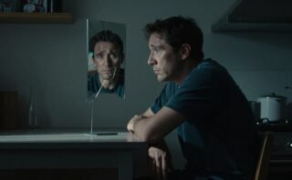 A worried man sits beneath a dim light beside a cracked mirror.